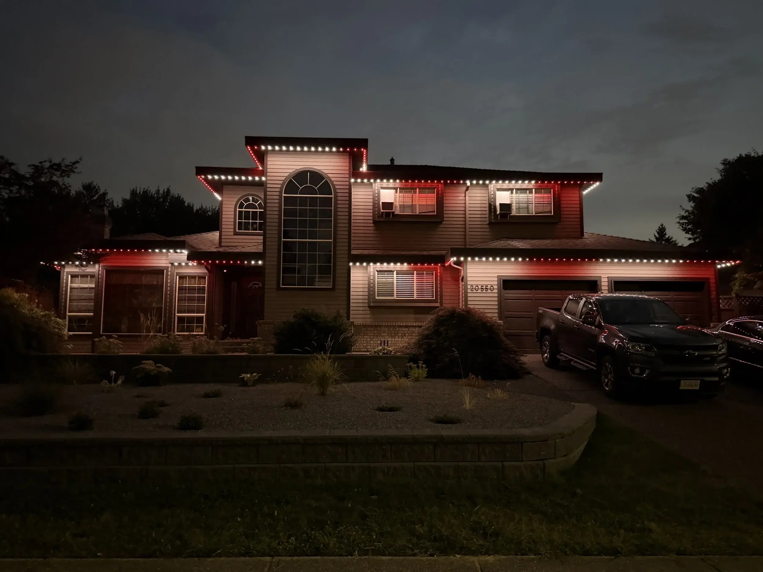 Two-story home featuring red and white permanent outdoor lighting at dusk