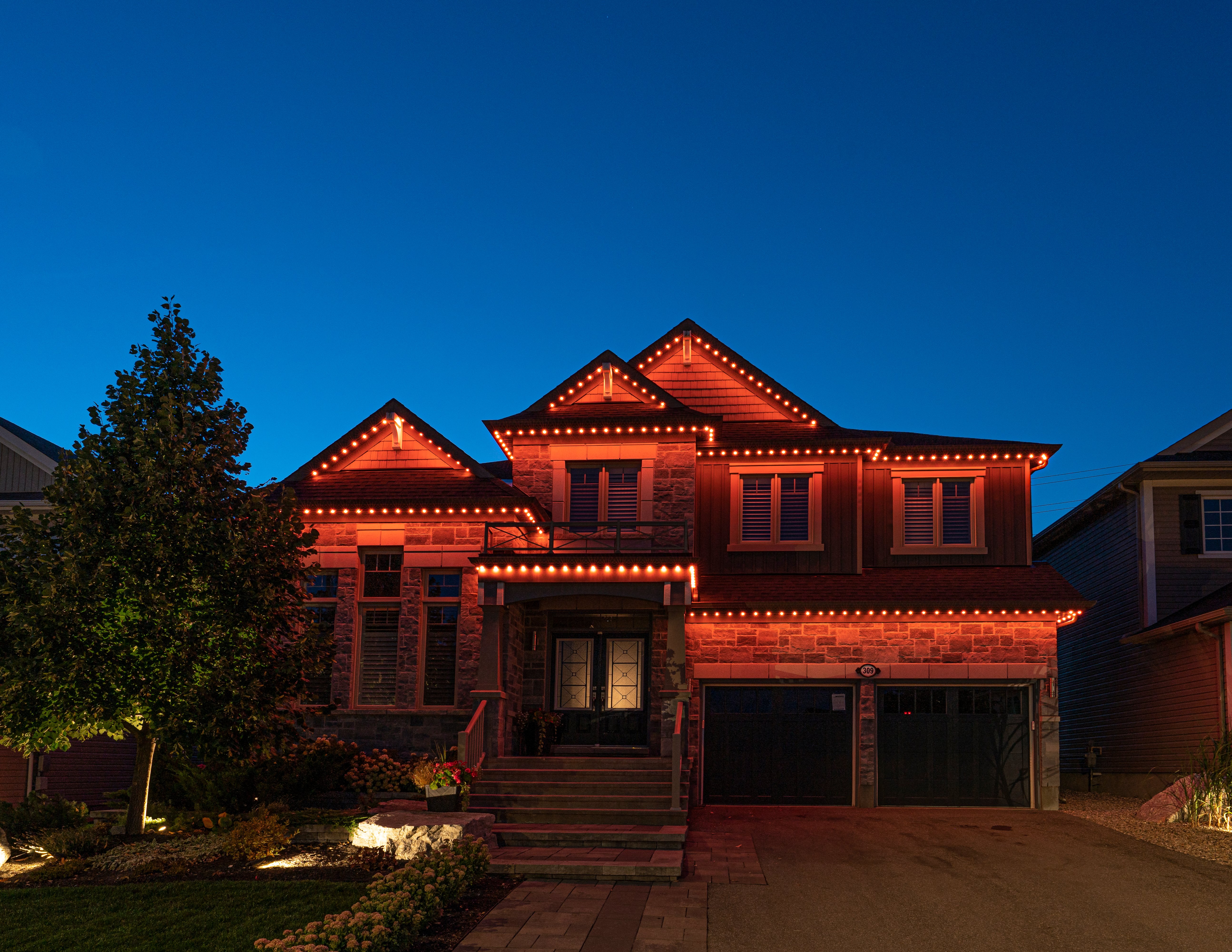 Two-story brick home with warm orange permanent outdoor lighting installed along the roofline at dusk