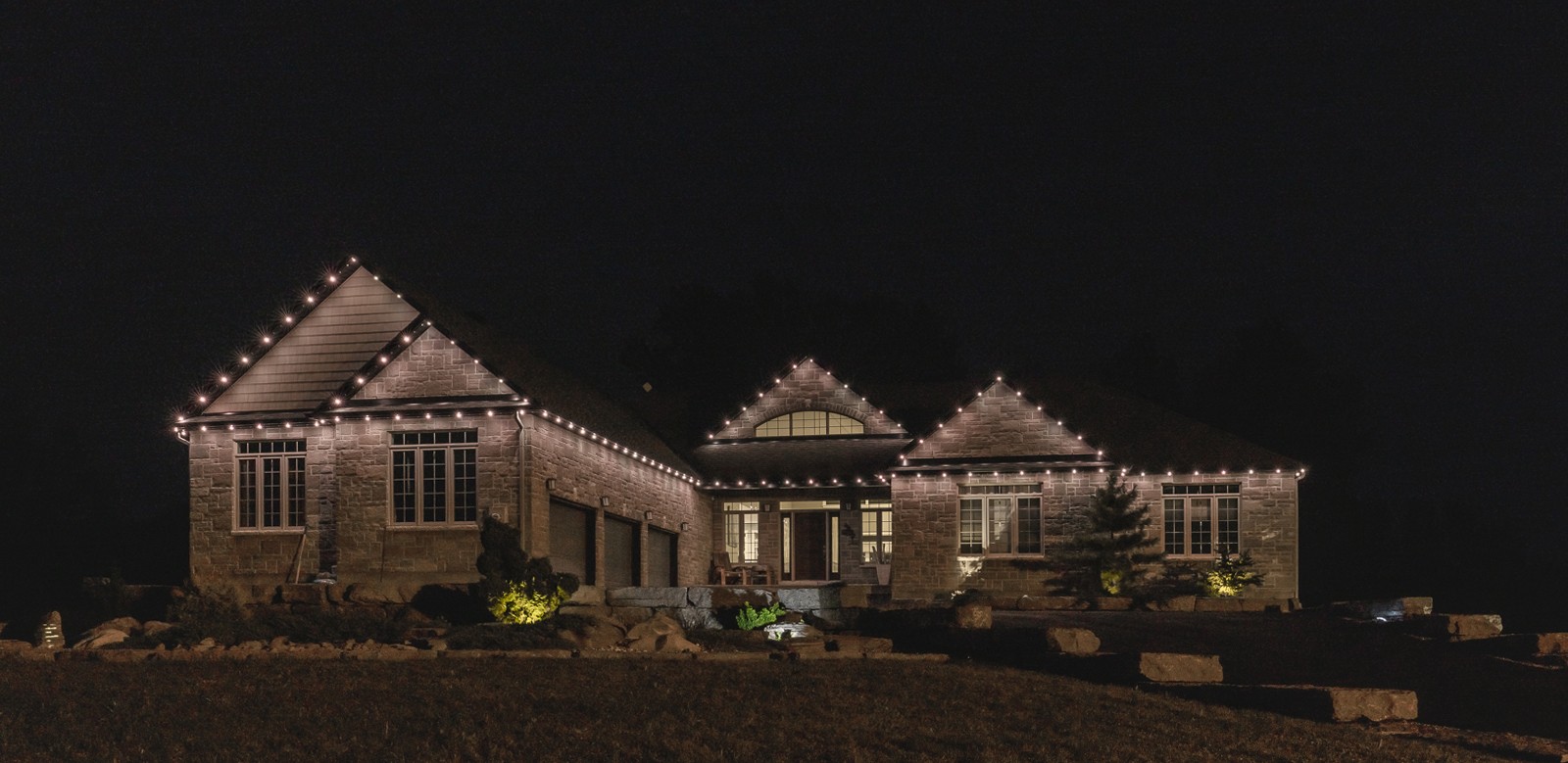 Brick house at night with permanent holiday lighting on the roofline.