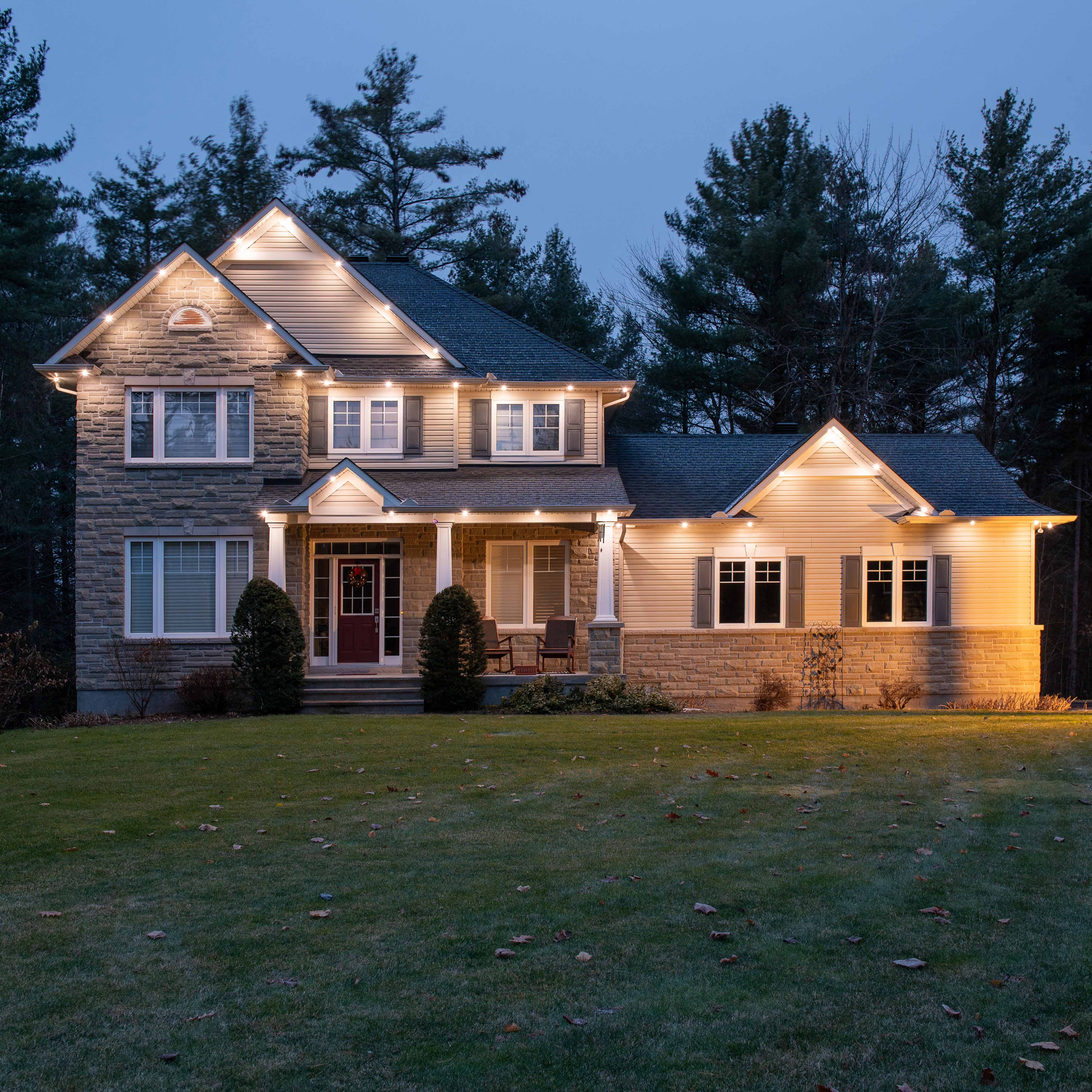 Festive red, green, and white permanent Christmas lights illuminating the house at dusk.