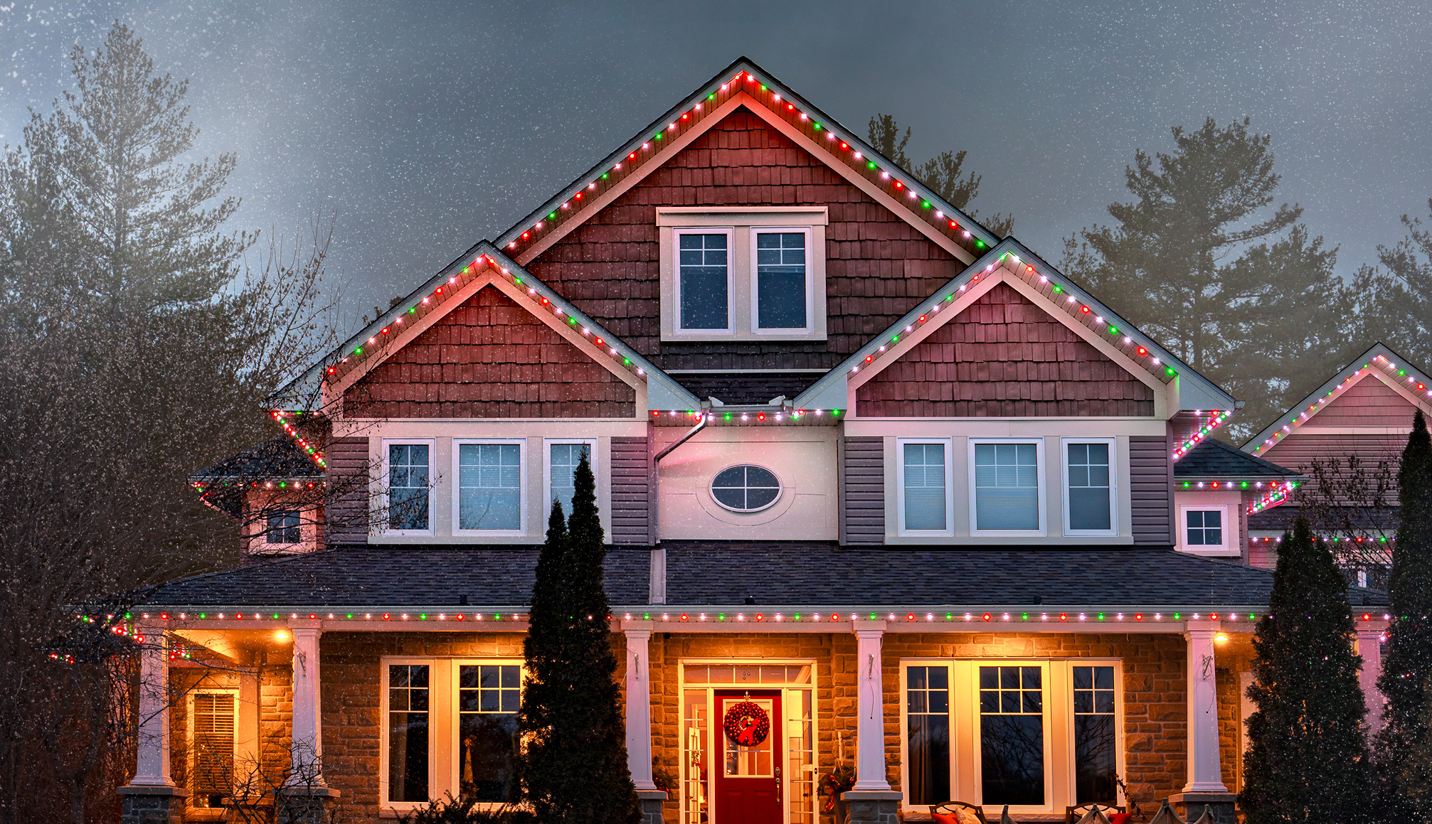 Red, green, and white permanent Christmas lights outlining the roofline of a house in a festive winter scene.