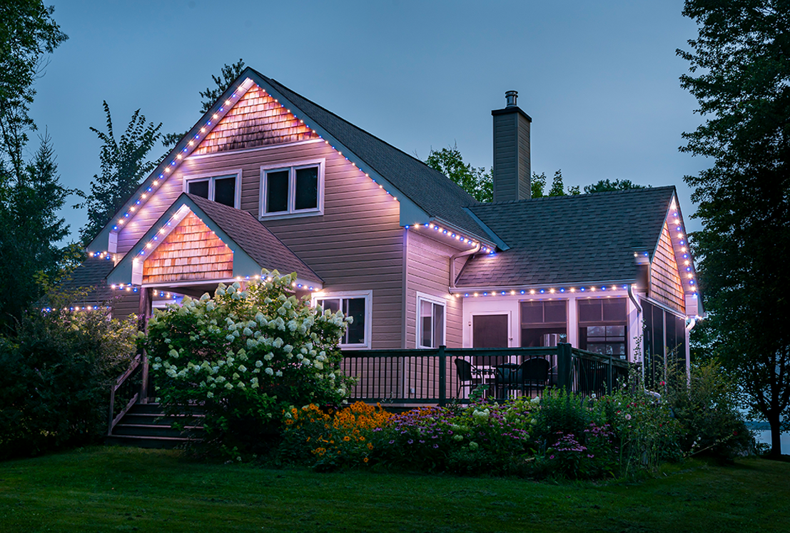 Permanent holiday lights in blue and white illuminating the roofline of a cottage at dusk.