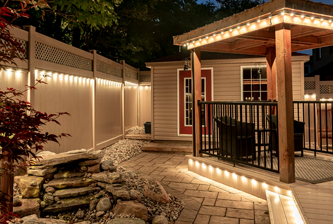 Warm white permanent lights illuminating a backyard fence, pergola, and patio area in the evening.