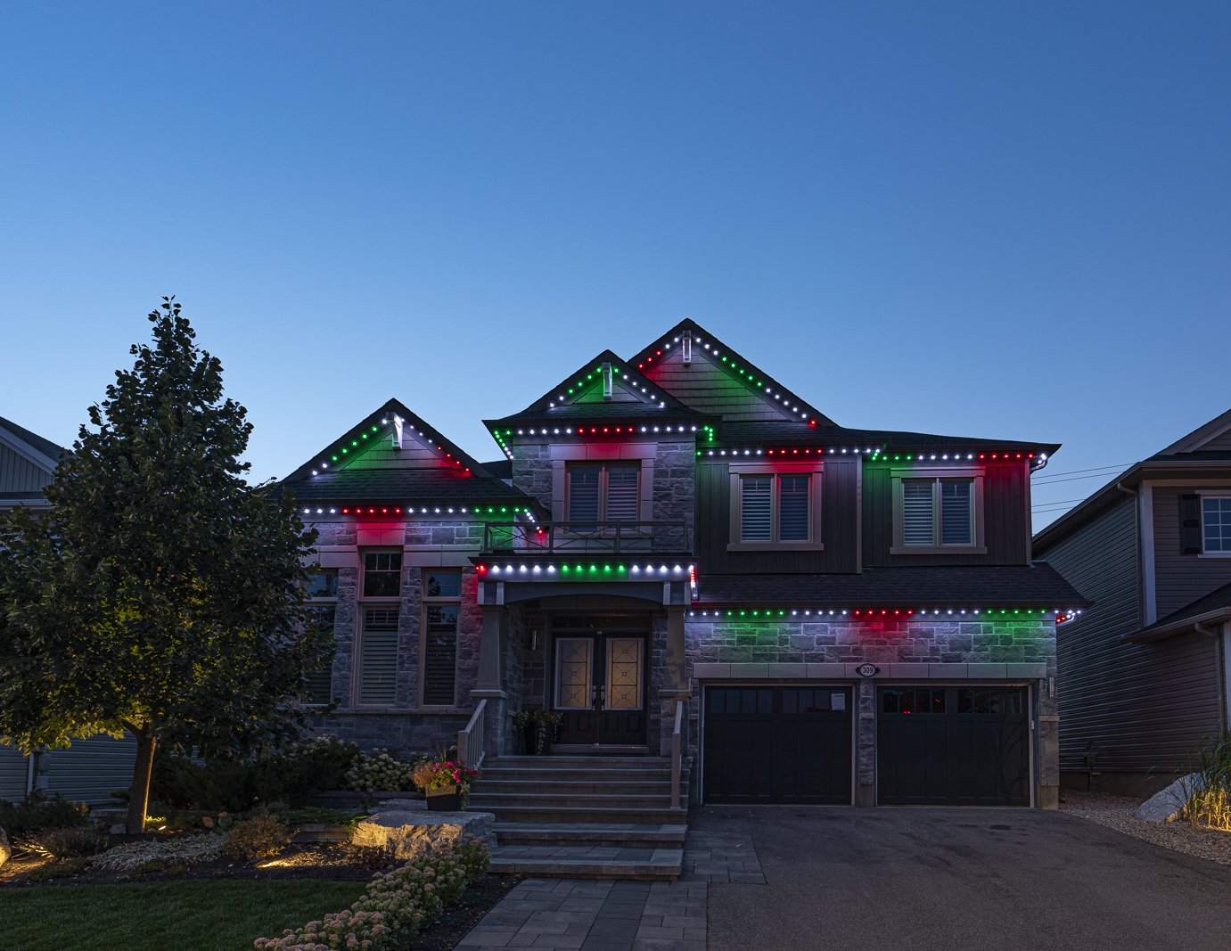 Festive red, green, and white permanent Christmas lights illuminating the house at dusk.