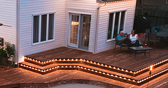 Deck outlined with warm permanent outdoor lights at dusk, with two people seated nearby.