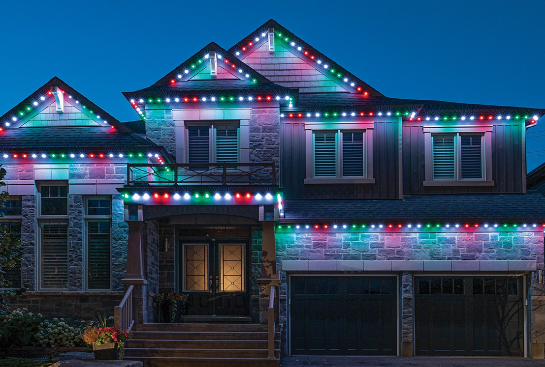 House outlined with permanent Christmas lights in red, green, and white glowing along the roofline at dusk.