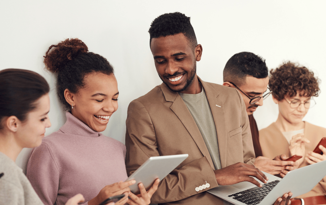 A group of people smiling and using a tablet, laptop, and phones together.