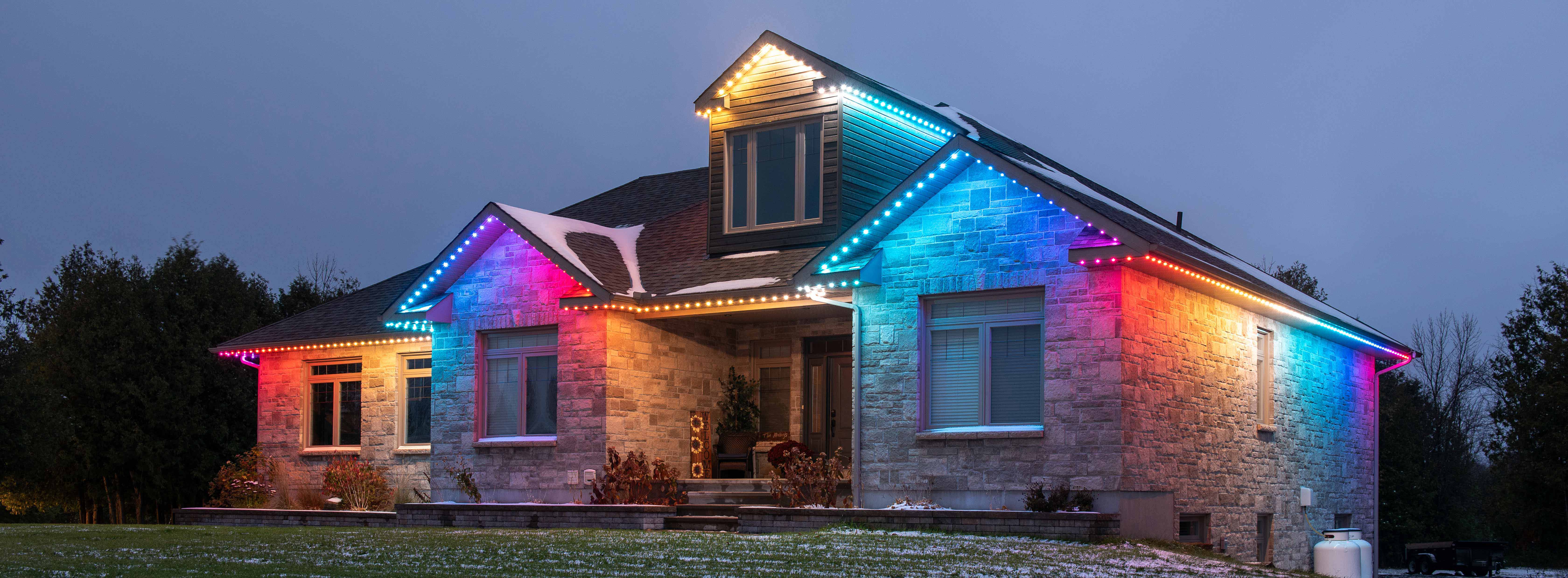 Stone home at dusk illuminated with permanent holiday lighting along the roofline and gables.