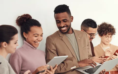 A group of people smiling and using a tablet, laptop, and phones together.