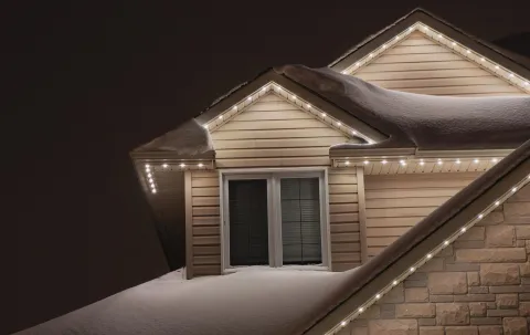 Snow-covered house roof at night with permanent holiday lighting installed along the eaves.